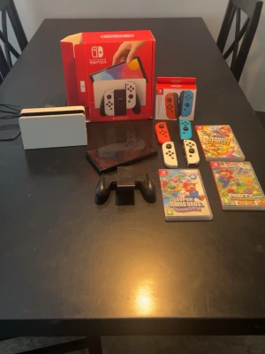 Nintendo Switch OLED med extra Joy-Cons - Nintendo Switch OLED med vit docka, två par Joy-Con-kontroller (ett vitt och ett röd/blått), handkontrollhållare och tre spel: Super Mario Bros. Wonder, Mario Party Superstars och Mario Party Jamboree. Perfekt för gamingkvällar och multiplayer.