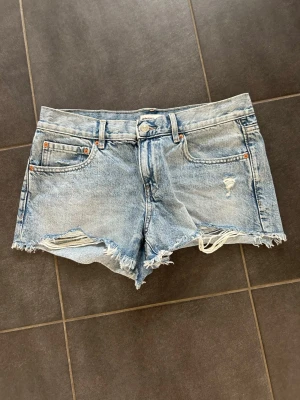 Ljusblå denimshorts med fransig kant - Säljer ett par ljusblå denimshorts med fransig kant och slitna detaljer. Klassisk femficksmodell med bälteshällor och knappgylf. Perfekta till sommaren för en avslappnad och trendig look.