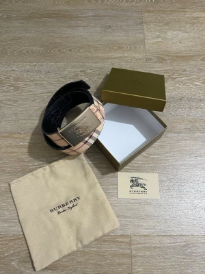Beige rutigt bälte - Snyggt bälte i klassiskt beige och brunt rutigt mönster med stor guldfärgad metallspänne. Bältet har exklusiv känsla och kommer med originalpåse och box. Perfekt accessoar för att lyfta din outfit.