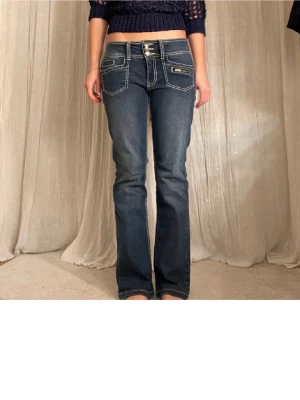 Mörkblå bootcut jeans med kontrastsömmar - Snygga mörkblå bootcut jeans med vita kontrastsömmar och tre knappar i midjan. Jeansen har flera fickor, inklusive en med dragkedja fram, och en klassisk tvättad look. Perfekta för dig som gillar en avslappnad men trendig stil.
