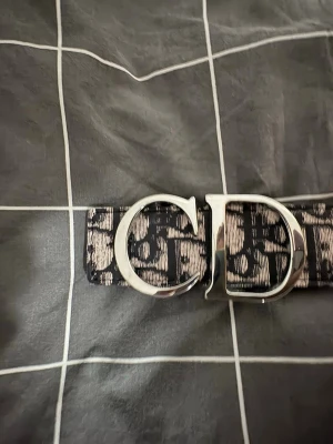 Svart och vit bälte CD logga  - Snyggt svart och vitt bälte med CD märke på. Bältet har ett mönster med bokstäver över hela skärpet. Perfekt accessoar för att lyfta din outfit och ge den en exklusiv touch. Den är storlek 110cm.