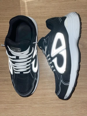 Svarta sneakers med vita detaljer - Snygga svarta sneakers med vita reflekterande detaljer och vita snören. Skorna har en sportig design med tjock vit sula och meshpartier för extra komfort. Perfekta för dig som gillar streetwear och vill sticka ut med coola detaljer.