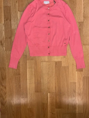 Rosa cardigan med knappar - Söt rosa cardigan med rund halsringning och långa ärmar. Tröjan har knäppning framtill med små metallknappar och ribbade muddar vid ärmslut och nederkant. Perfekt att slänga över en topp för en färgklick i vardagen.