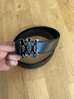 Svart bälte med metallspänne - Snyggt svart bälte i skinn med ett stort, unikt metallspänne som har en cool design. Perfekt för att ge din outfit en edgy touch. Bältet är stilrent och passar till många olika looks.