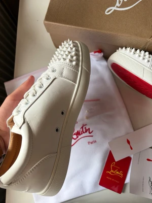 Vita Louboutin sneakers med nitar - Säljer ett par vita sneakers från Christian Louboutin med ikoniska röda sulor och coola nitar på tån. Skorna är låga och har en stilren design i läder med vita snören. Perfekta för dig som vill sticka ut med exklusiva detaljer och streetstyle-vibe.