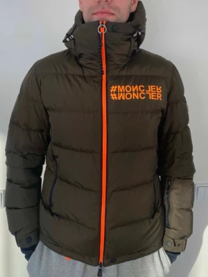 Olivgrön pufferjacka med orange detaljer - Säljer en olivgrön pufferjacka med orange dragkedja och tryck på bröstet. Jackan har hög krage, huva med dragsko och flera fickor med svarta dragkedjor. Perfekt för kalla vinterdagar och riktigt snygg streetstil. In very good condition. Moncler ISORNO Giubbotto. Size 2. Bought in 2022. 100% authentic, bought for 2100 euros.💶 