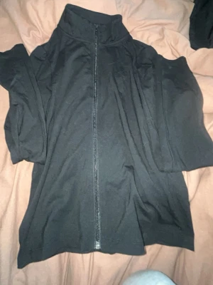 Svart zip-hoodie med hög krage - En svart hoodie med dragkedja framtill och hög krage. Modellen är enkel och stilren, perfekt för en chill look. Tillverkad i mjukt material som känns skönt mot huden. Passar dig som gillar basic och bekvämt.