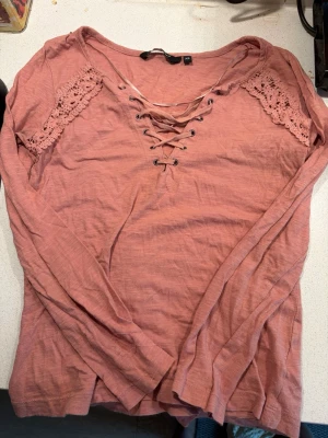 Rosa långärmad topp med snörning - Söt långärmad topp i dammigt rosa med snörning framtill och virkade spetsdetaljer vid axlarna. Toppen har en v-ringad hals och är gjord i mjukt bomullstyg. Perfekt för dig som gillar romantiska detaljer och en avslappnad stil.