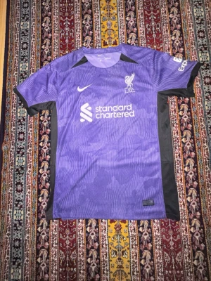 Lila Liverpool FC matchtröja Nike - Säljer en lila Liverpool FC matchtröja från Nike med vita detaljer och klubbmärke på bröstet. Tröjan har svarta inslag på sidorna och axlarna samt tryckt sponsorlogga framtill. Perfekt för dig som älskar fotboll och vill stötta ditt lag.