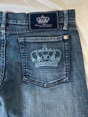 Blå bootcut jeans Victoria Beckham - Snygga blå bootcut jeans från Victoria Beckham med ljus tvätt och broderad krona på bakfickan. Klassisk femficksmodell med knappgylf och coola slitningar. Perfekta för dig som gillar en avslappnad men trendig look.