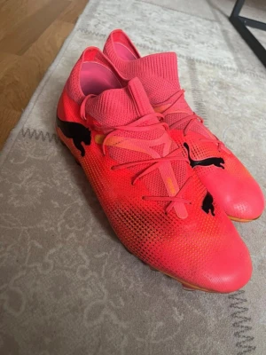 Rosa fotbollsskor från Puma - Snygga fotbollsskor från Puma i en stark rosa färg med svarta Puma-loggor på sidan och ovansidan. Skorna har strumpliknande skaft, snörning och ovandel i mesh-material för bra ventilation och passform. Perfekta för dig som vill sticka ut på planen.