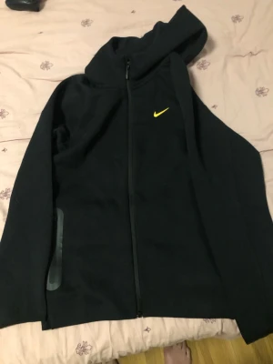 Nocta tracksuit  - Säljer en svart hoodie från Nike med dragkedja framtill och gul Nike-logga på bröstet. Hoodien har huva och långa ärmar, samt en diskret ficka på sidan. Perfekt för en sportig och avslappnad stil.