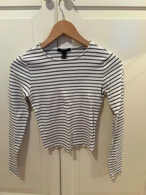 Randig långärmad topp - Säljer en vit långärmad topp från Forever 21 med svarta horisontella ränder. Toppen har en rund halsringning och är i en slim passform. Perfekt att styla med jeans eller kjol för en enkel och snygg look.