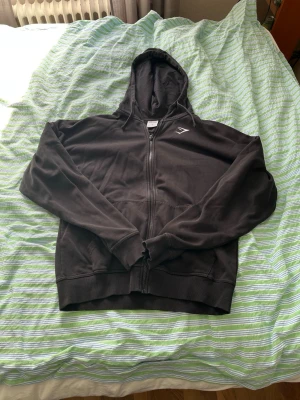 Svart zip hoodie från Gymshark - Säljer en svart hoodie från Gymshark med dragkedja framtill och huva. Hoodien har två fickor och en liten Gymshark-logga på bröstet. Tillverkad i mjukt material som passar perfekt till chill eller träning.