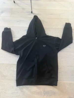 Svart hoodie med hjärta och ögon - Säljer en svart hoodie med dragkedja och huva. På bröstet finns ett litet hjärta med ögon och på ryggen ett stort hjärta med ögonmotiv. Hoodien har fickor framtill och ribbade muddar. Perfekt för dig som gillar streetwear och coola prints.