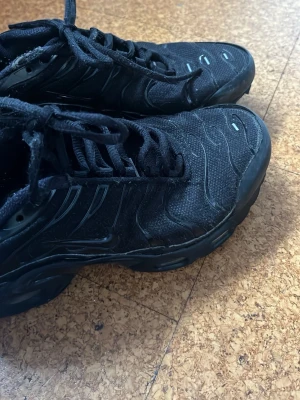 Svarta Tn Nike skor  - Säljer ett par svarta Nike Air Max Plus sneakers med klassisk vågig design och mesh-överdel. Skorna har svarta snören, diskreta swoosh-loggor och detaljer i gult och rött på sulan. Perfekta för dig som gillar streetwear och sportig stil.