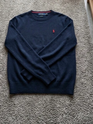 Mörkblå stickad tröja Polo Ralph Lauren - Mörkblå stickad tröja från Polo Ralph Lauren med klassisk röd logga broderad på bröstet. Tröjan har rund halsringning, ribbade muddar och är långärmad. Perfekt för dig som gillar stilrena och tidlösa plagg.