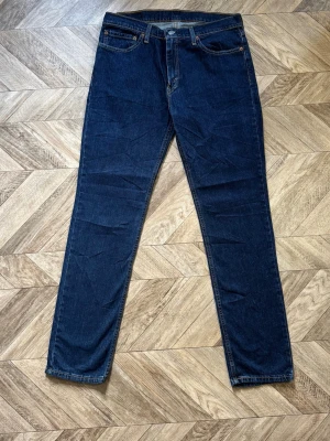 Mörkblåa Levi's 511 - Säljer ett par klassiska mörkblå jeans från Levi's med rak passform och fem fickor. Jeansen har en tidlös look med normal midja och Levi's ikoniska läderpatch bak i linningen. Perfekta till alla tillfällen och lätta att matcha.Längd 109cm Bredd 41cm