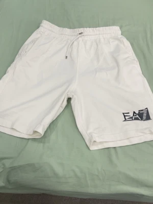 Vita EA7 shorts med snörning - Säljer ett par vita shorts från EA7 Emporio Armani med svart logga på vänster ben. Shortsen har elastisk midja med snörning och sidofickor. Perfekta för sommaren och har en clean, sportig look.