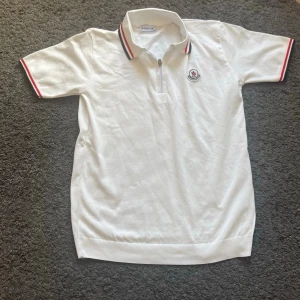 Moncler polotopp - Säljer en vit polotopp från Moncler med korta ärmar och dragkedja framtill. Kragen har röd och marinblå randig detalj och Moncler-logga är broderad på bröstet. Perfekt för dig som gillar klassisk och sportig stil. Priset kan diskuteras 