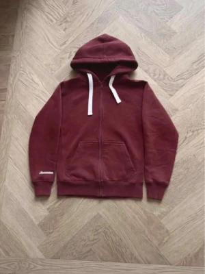 Vinröd hoodie med dragkedja - Säljer en vinröd hoodie med dragkedja och vita snören i huvan. Hoodien har en klassisk känguruficka framtill och ribbade muddar vid ärmar och nederkant. Perfekt för en avslappnad stil och enkel att matcha med jeans eller joggers.