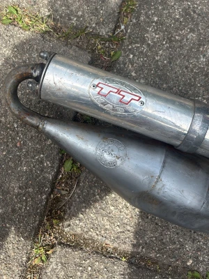 TT avgassystem Leovince Speedfight 2 - Säljer ett TT avgassystem från Leovince i metall, perfekt för mopeder eller mindre motorfordon. Systemet är i silverfärgat stål och har en klassisk böjd form med tydliga logotyper på ljuddämparen och expansionskammaren.