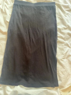 Svart ribbad långkjol med slits - Säljer en svart långkjol i ribbad bomull med en snygg slits på sidan. Kjolen har en enkel och stilren design som passar perfekt till många olika outfits. Superskön och lätt att matcha!