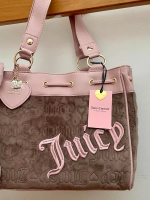 Juicy Couture rosa och brun handväska daydreamer  - Säljer en ikonisk handväska från Juicy Couture i mjuk brun velour med ljusrosa detaljer och broderad logga. Väskan har guldfärgade metalldetaljer, hjärtformad tag och breda axelband. Perfekt för dig som gillar Y2K-stil och vill sticka ut.