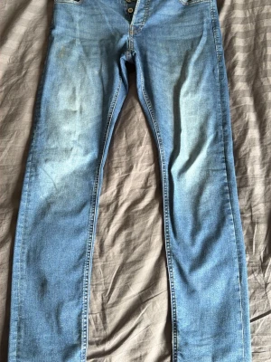 Ljusblå raka jeans  - Säljer ett par ljusblå jeans med raka ben och knappgylf. Jeansen har klassisk femficksdesign och slitningar framtill för en avslappnad look. Materialet är denim i bomull och passformen är normal. Perfekta för en casual stil.