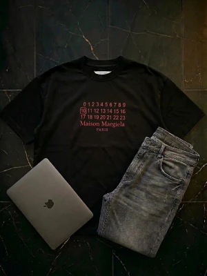 Maison Margiela svart t-shirt med rött tryck - Maison Margiela svart t-shirt med tryck i rött framtill. Klassisk rund hals och korta ärmar. Snygg och stilren design med Maison Margiela Paris-logga. Perfekt för dig som gillar exklusiva streetwear-plagg. Tröjan är helt oanvänd och är perfekt inför skolstarten. Tröjan är markerad med storleken 48 men passar storleken L.