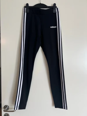 Svarta leggings med vita ränder Adidas - Snygga svarta leggings från Adidas med klassiska vita ränder längs benen och Adidas-logga på låret. Stretchigt material och smal passform som sitter tajt. Perfekta för en sportig och avslappnad stil. Är i en barnstorlek men passar också vuxna som är max 164 cm långa.   Midja: ca 32 cm Längd: ca 85 cm