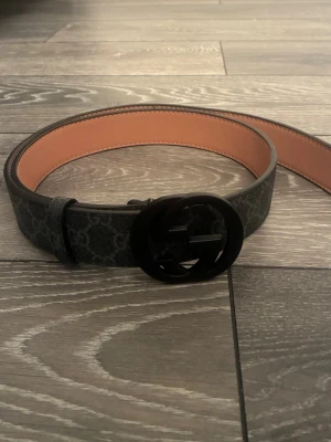 Gucci bälte - Snyggt svart skärp från Gucci med klassiskt GG-mönster och rund, svart spänne med GG-logga. Insidan är beige och materialet är skinn. Perfekt accessoar för att lyfta din outfit.
