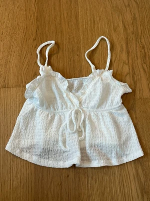 Vit croppad linnetopp med knyt - Söt vit croppad linnetopp med tunna axelband och knyt framtill. Toppen har en lätt rynkad struktur och v-ringad hals. Perfekt för varma dagar och enkel att styla med jeans eller kjol.