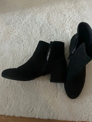 Svarta stövletter med klack - Snygga svarta stövletter i mockaimitation med dragkedja på sidan och rundad tå. Skorna har en stabil blockklack som ger extra höjd och stil. Perfekta för dig som vill ha en klassisk och trendig look till hösten eller vintern. Bara använda en gång 💕 butikspriset 499:-
