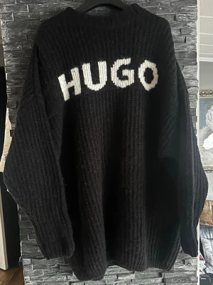 Svart stickad tröja med HUGO-tryck - Cool svart stickad tröja från HUGO med vit text över bröstet. Oversized passform, ribbstickad struktur och rund halsringning. Perfekt för dig som gillar streetstyle och vill ha något bekvämt och trendigt.
