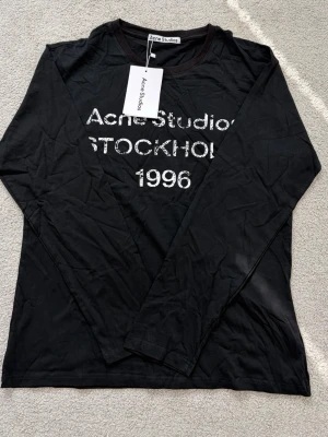 Svart långärmad topp Acne Studios - Svart långärmad topp från Acne Studios med tryck i vitt framtill där det står 'Acne Studios STOCKHOLM 1996'. Rund halsringning och mjukt material som känns skönt mot huden. Perfekt för en cool och avslappnad stil.