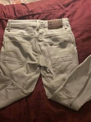 Gstar jeans ljusgråa  - Säljer ett par ljusgrå jeans med raka ben och klassisk femficksdesign. Jeansen har bälteshällor, knappgylf och detaljerade sömmar på bakfickorna. Materialet ser ut att vara bomullsdenim med en mjuk finish.