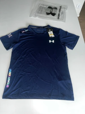 Blå tränings-tshirt från Under Armour - Snygg mörkblå tränings-tshirt från Under Armour med korta ärmar och rund hals. Logga på bröstet och färgglad text längs sidan. Tillverkad i ett lätt och snabbtorkande funktionsmaterial som passar perfekt till träning. 