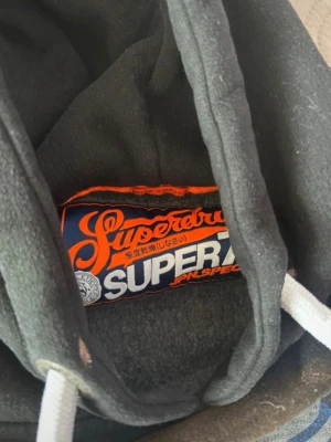 Svart hoodie från Superdry - Snygg svart hoodie från Superdry med stor logga i blått och vitt på bröstet. Klassisk huva med vita dragsnören och mjukt material på insidan. Perfekt för en avslappnad och cool stil.