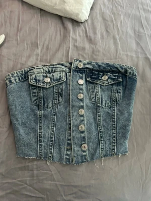 Blå denim tubtopp med knappar - Säljer en snygg blå denim tubtopp med knappar framtill och två dekorativa bröstfickor. Toppen har rå kant nertill och smockad rygg för skön passform. Perfekt till sommarens outfits och lätt att styla med olika accessoarer.