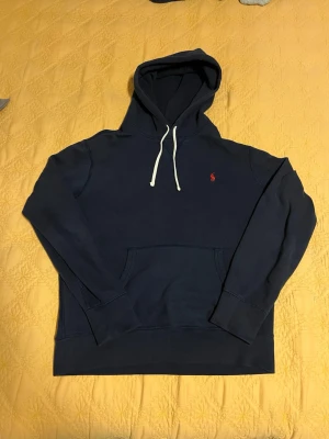 Mörkblå hoodie från Polo Ralph Lauren - Mörkblå hoodie från Polo Ralph Lauren med klassisk röd logga broderad på bröstet. Hoodien har vit dragsko i huvan och en stor känguruficka framtill. Tillverkad i mjukt bomullsmaterial och har ribbade muddar vid ärmslut och nederkant.