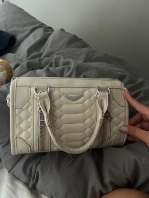 Beige quiltad handväska med detaljer - Snygg beige handväska i quiltat mönster med två handtag och silverfärgade metalldetaljer. Väskan har två dragkedjor framtill och en liten metallskylt med dekorativt mönster. Tillverkad i konstläder och har en kompakt, boxig form.