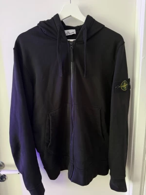 Svart hoodie från Stone Island - Säljer en svart hoodie från Stone Island med dragkedja framtill, huva med dragsko och känguruficka. Klassisk Stone Island-patch på vänster ärm. Hoodien är i mjukt bomullsmaterial och har en clean, stilren look som passar perfekt till streetwear.