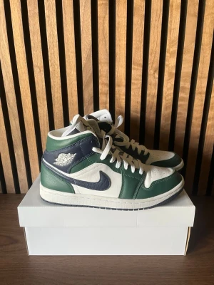 Nike Air Jordan 1 Mid grön/vit - Snygga Nike Air Jordan 1 Mid sneakers i grönt, vitt och mörkblått. Klassisk siluett med högt skaft, vita snören och ikoniska Swoosh-loggan på sidan. Tillverkade i läder med perforerad tå för extra komfort.