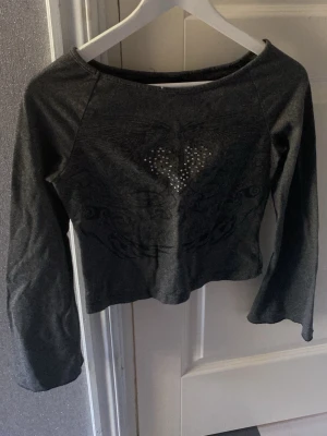 Mörkgrå croppad topp med hjärtdetalj - Snygg mörkgrå croppad topp med lång ärm och ett hjärtmotiv framtill dekorerat med små glittriga stenar. Toppen har en tight passform och är perfekt för dig som gillar edgy stil.