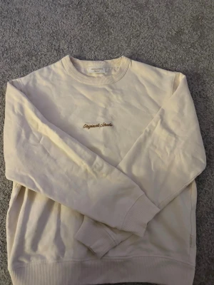 Beige sweatshirt Originals Studio - Beige sweatshirt med rund hals och ribbade muddar. På bröstet finns en broderad text i brunt där det står 'Originals Studio'. Tröjan har en avslappnad passform och är perfekt för en clean och enkel stil.