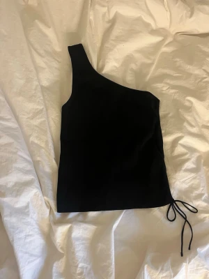 Svart oneshoulder topp med knyt - Snygg svart oneshoulder topp med knyt i sidan. Toppen har en asymmetrisk axel och är tillverkad i ett mjukt material som sitter tight mot kroppen. Perfekt för dig som vill ha en stilren och trendig look.