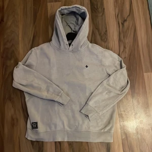Ljusgrå hoodie från Morris Stockholm - Säljer en stilren ljusgrå hoodie från Morris Stockholm med broderad logga på bröstet och patch nertill. Tröjan har huva, långa ärmar och är tillverkad i mjuk bomullsmix. Perfekt för en chill och snygg look.