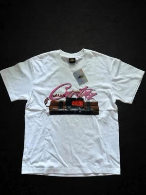 Vit t-shirt med tryck från Adieuite - Vit t-shirt från Adieuite med ett stort grafiskt tryck framtill som visar en digital bomb och rosa graffiti-text. Klassisk passform med rund hals och korta ärmar. Tillverkad i mjuk bomull, perfekt för en streetwear-look.