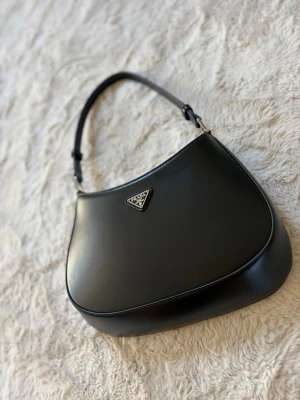 Svart axelremsväska  - Snygg svart axelremsväska från i slätt läder med ikonisk metallogo framtill. Väskan har en stilren, rundad form och ett rymligt innerfack med magnetknäppning. Perfekt accessoar för dig som gillar klassisk och trendig design. Mycket fint skick!